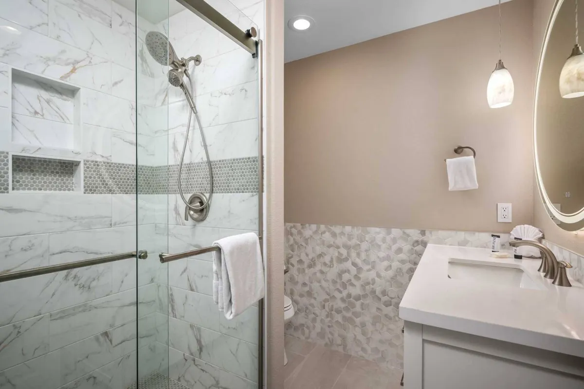 A sleek bathroom featuring a glass shower enclosure, marble tile surfaces, a contemporary vanity with sink, and pendant lighting. The space exudes a calm, serene, and modern vibe, evoking feelings of a high-end spa environment.