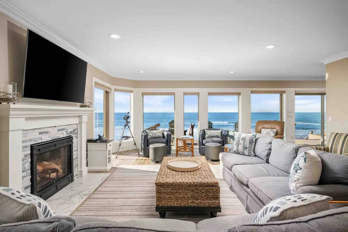The image showcases a cozy living room with a crackling fireplace, plush sofa, and expansive floor-to-ceiling windows offering breathtaking views of the Pacific Ocean. Warm sunlight fills the space as the ocean stretches out beyond, inviting relaxation and social gatherings against a backdrop of endless water and sky.