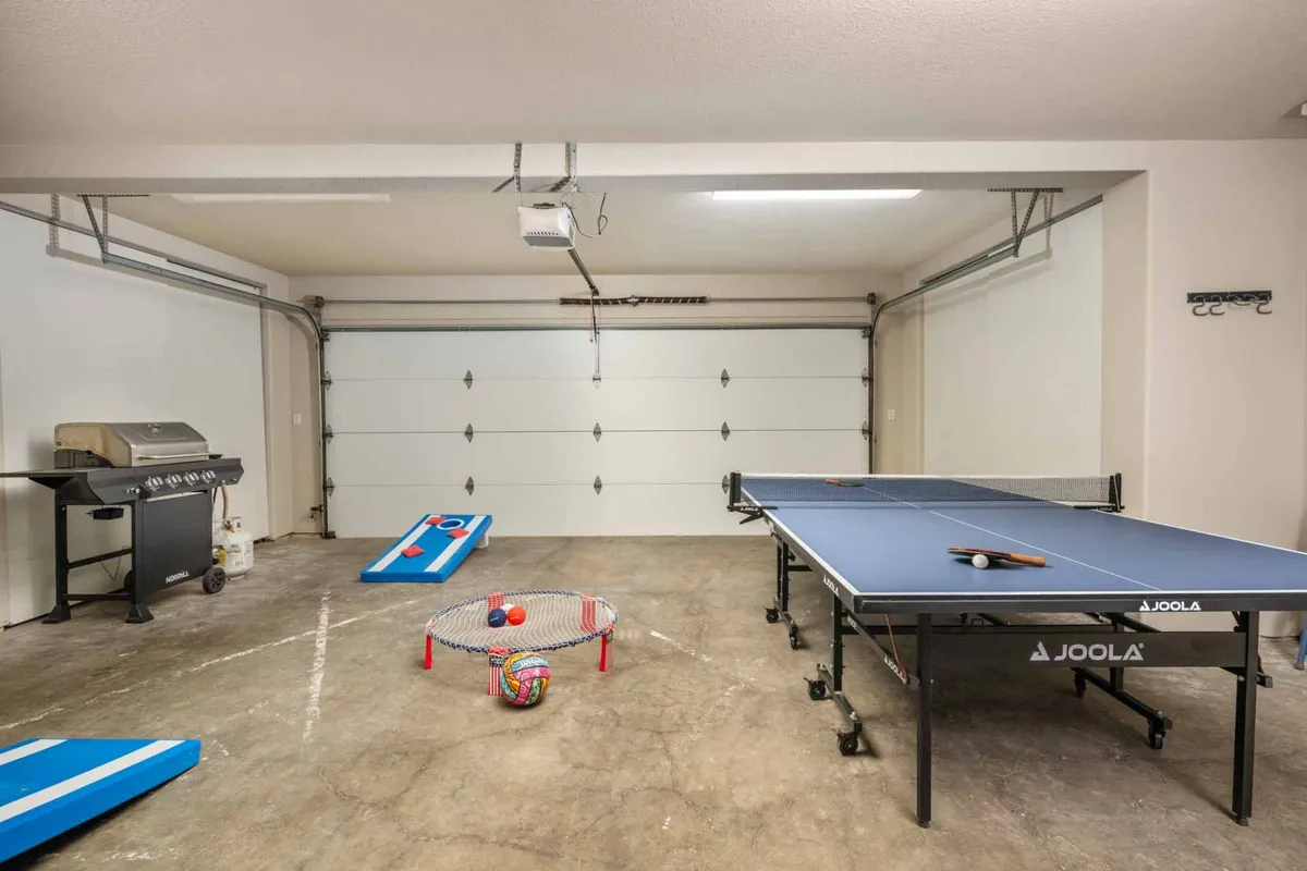 This image showcases a vibrant garage space featuring a ping pong table set up for play, a barbecue grill in the background, and a bean bag toss game with colorful balls, creating an inviting atmosphere for friendly competition and group fun.