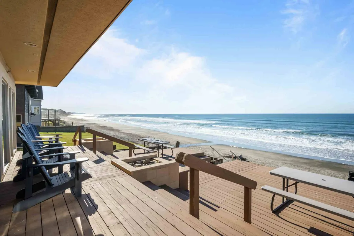 A view from a private deck overlooks the ocean, with comfortable deck seating and a fire pit in the foreground. The scene is relaxed, showing the ease of transitioning from indoor comfort to sandy beach, with clear opportunities for family gatherings, sandcastle building, and evenings by the fire as waves break nearby.