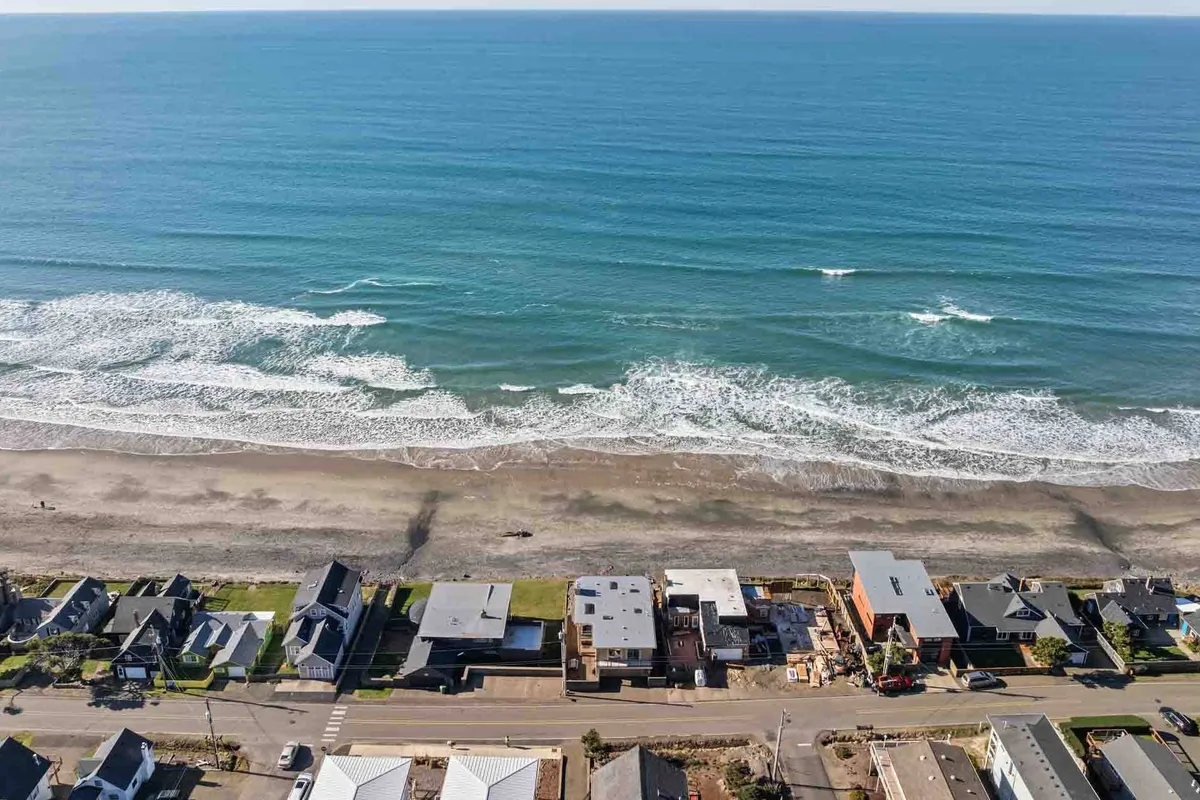 A wide-angle view featuring ocean waves, a line of beachfront homes, and a road running alongside the beach. People are seen walking along the shore, with children playing and building sandcastles. The scene is lively yet relaxed, embodying the communal and multi-generational enjoyment of the beach environment.