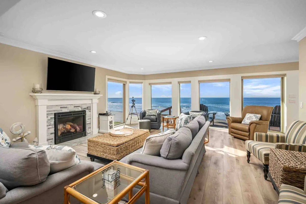 A spacious living area with a cozy seating arrangement faces a modern fireplace and a large television. Floor-to-ceiling windows reveal sweeping ocean views, flooding the room with natural coastal light and creating a seamless connection between the interior and the Pacific outside.