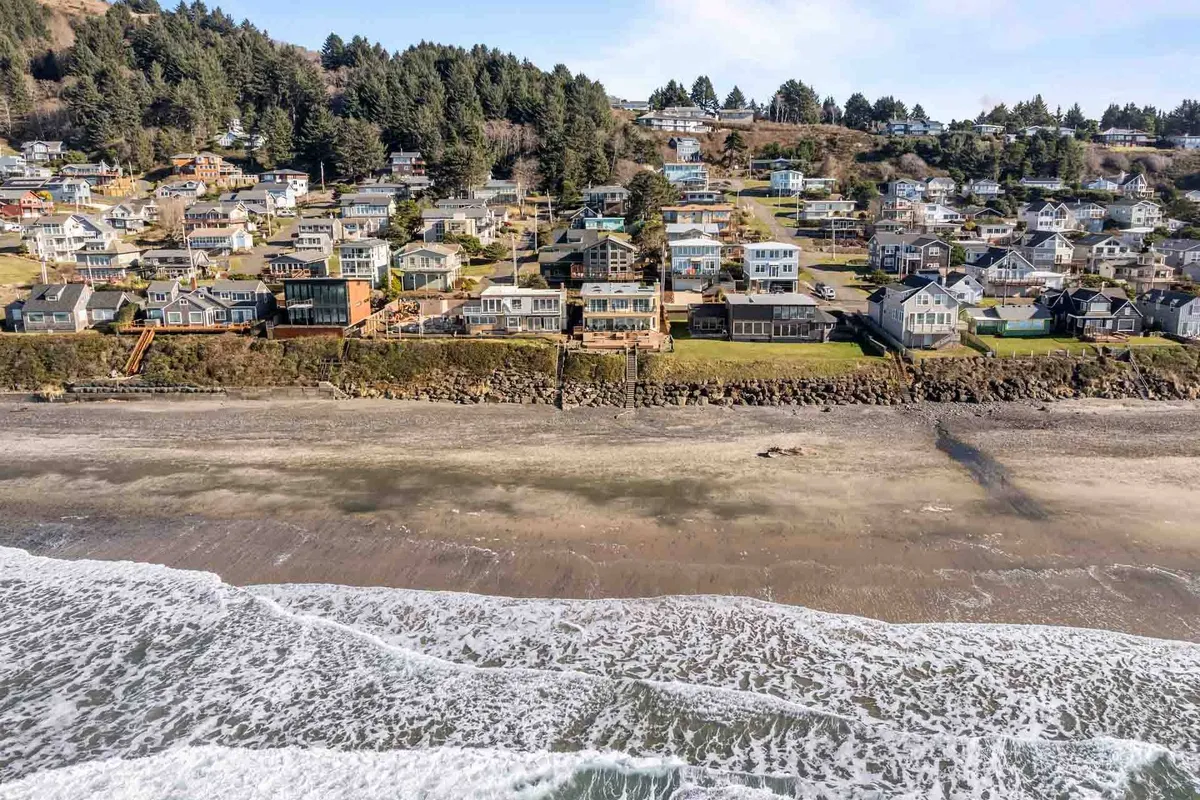 A panoramic exterior view captures classic beachfront houses framed by rolling green hills and waves crashing onto the shore. The scene evokes the essential Lincoln City vibe, with direct beach access and a sense of exploration evident in both the landscape and nearby homes.