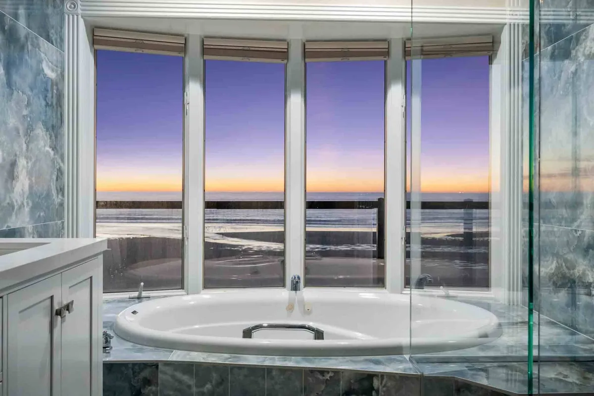 A modern bathroom with an oversized soaking tub positioned beneath large windows that reveal a sunset view. The abundance of natural light and tranquil setting evoke a spa-like ambiance, with the tub inviting relaxation and peaceful contemplation.