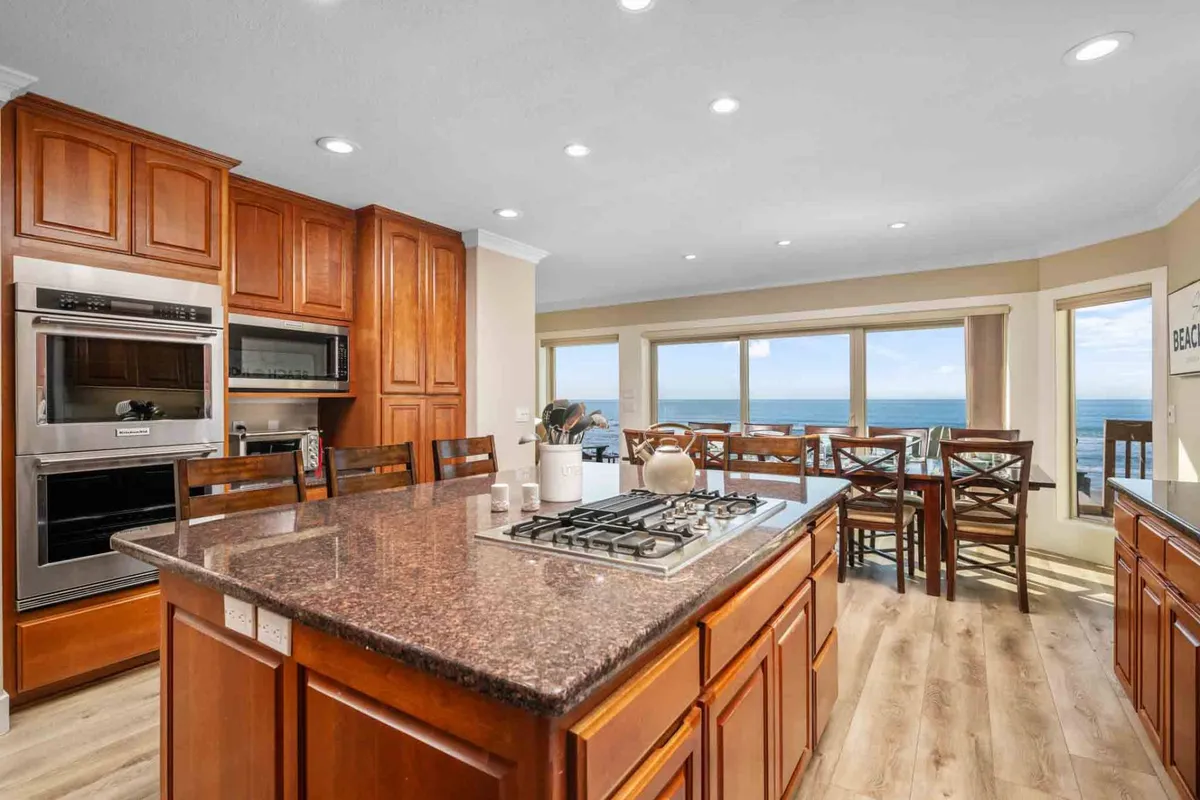 A welcoming open-concept kitchen and dining area featuring a granite island, wooden cabinetry, bar stools, and a large dining table surrounded by chairs. Expansive windows frame the spectacular ocean view, filling the space with light and highlighting the communal nature of the room.