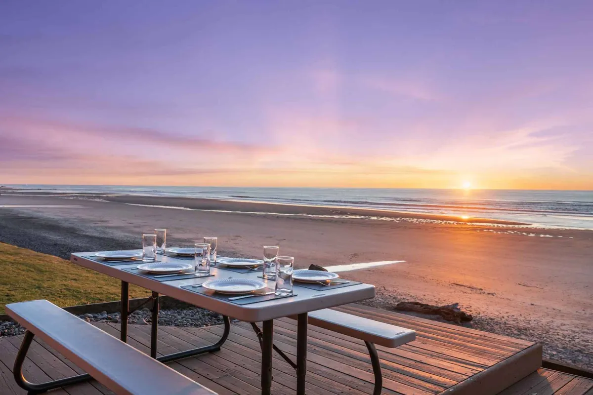 An inviting outdoor dining table is set for a meal, overlooking a sweeping ocean view as the sun sets. The scene highlights a tranquil patio space designed for gatherings, with the Pacific coastline serving as a dramatic backdrop. The setting exudes relaxation and the anticipation of shared meals and celebrations.