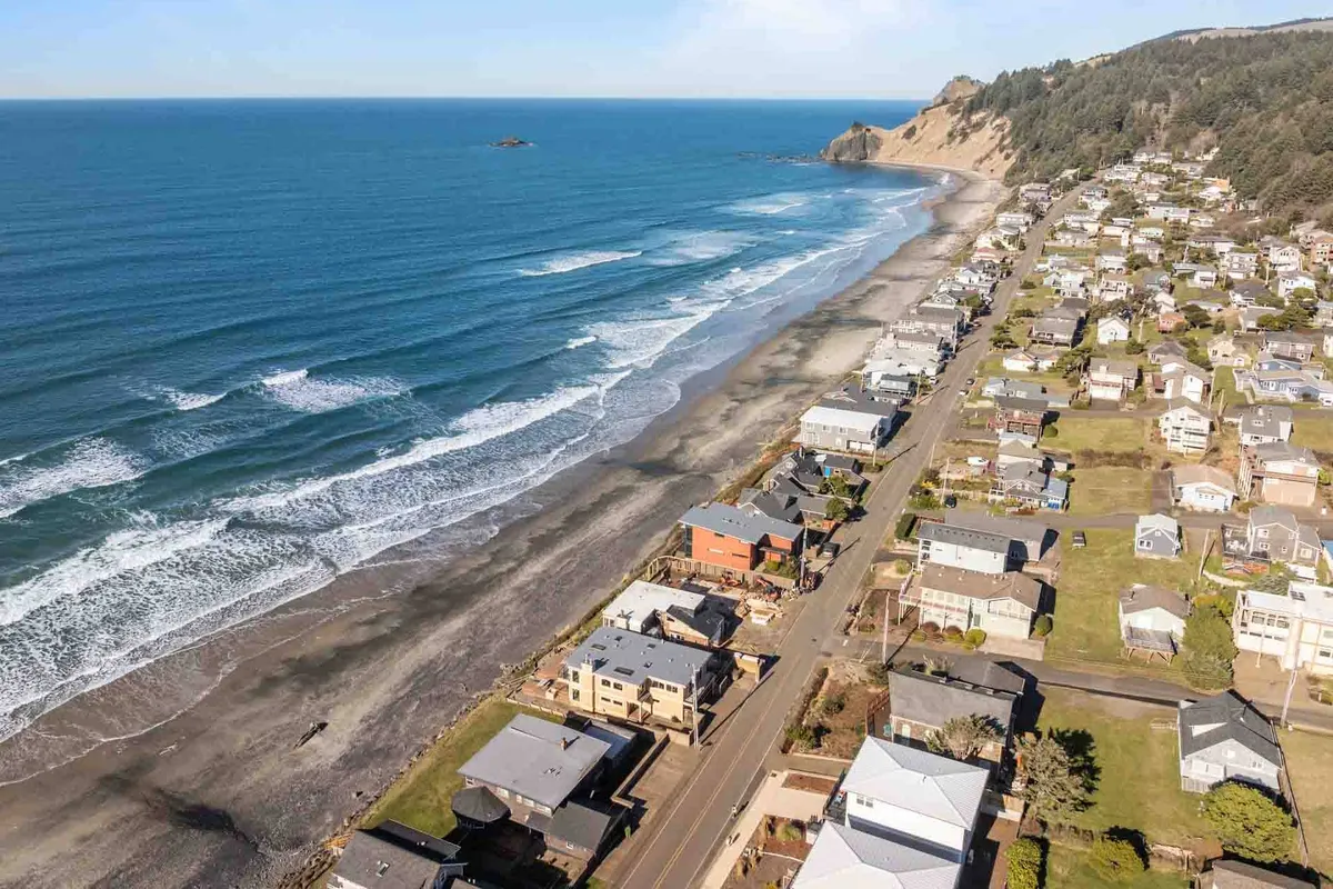 This image captures the coastline at Roads End, with gentle ocean waves meeting a wide sandy beach in front of charming beachfront houses. The scene shows people beachcombing and walking along the shore, evoking a lively, family-friendly atmosphere. The coastal landscape is expansive, with visible tide pools and rolling surf under a serene sky.