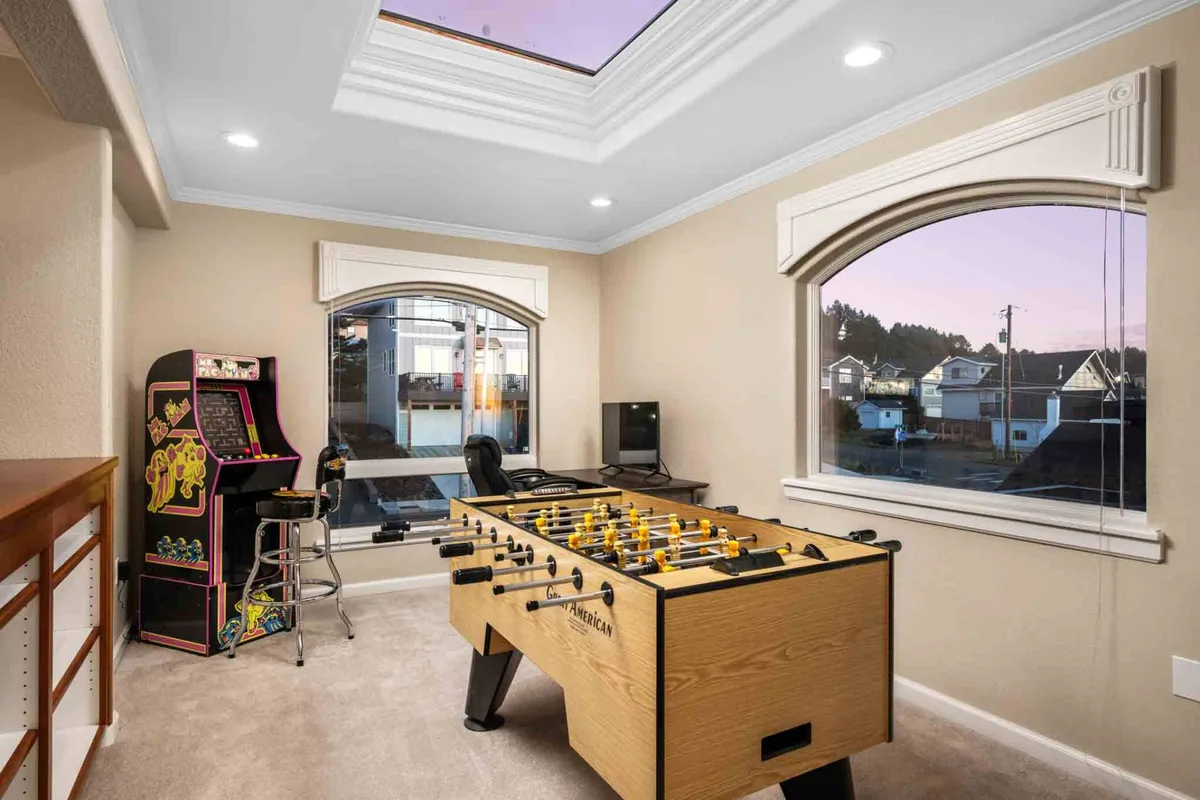 A bright, sunlit loft area filled with gaming options, including a foosball table and a classic arcade machine, with windows revealing glimpses of the outdoors. A work desk is also visible, emphasizing the space’s flexibility for both play and productivity.